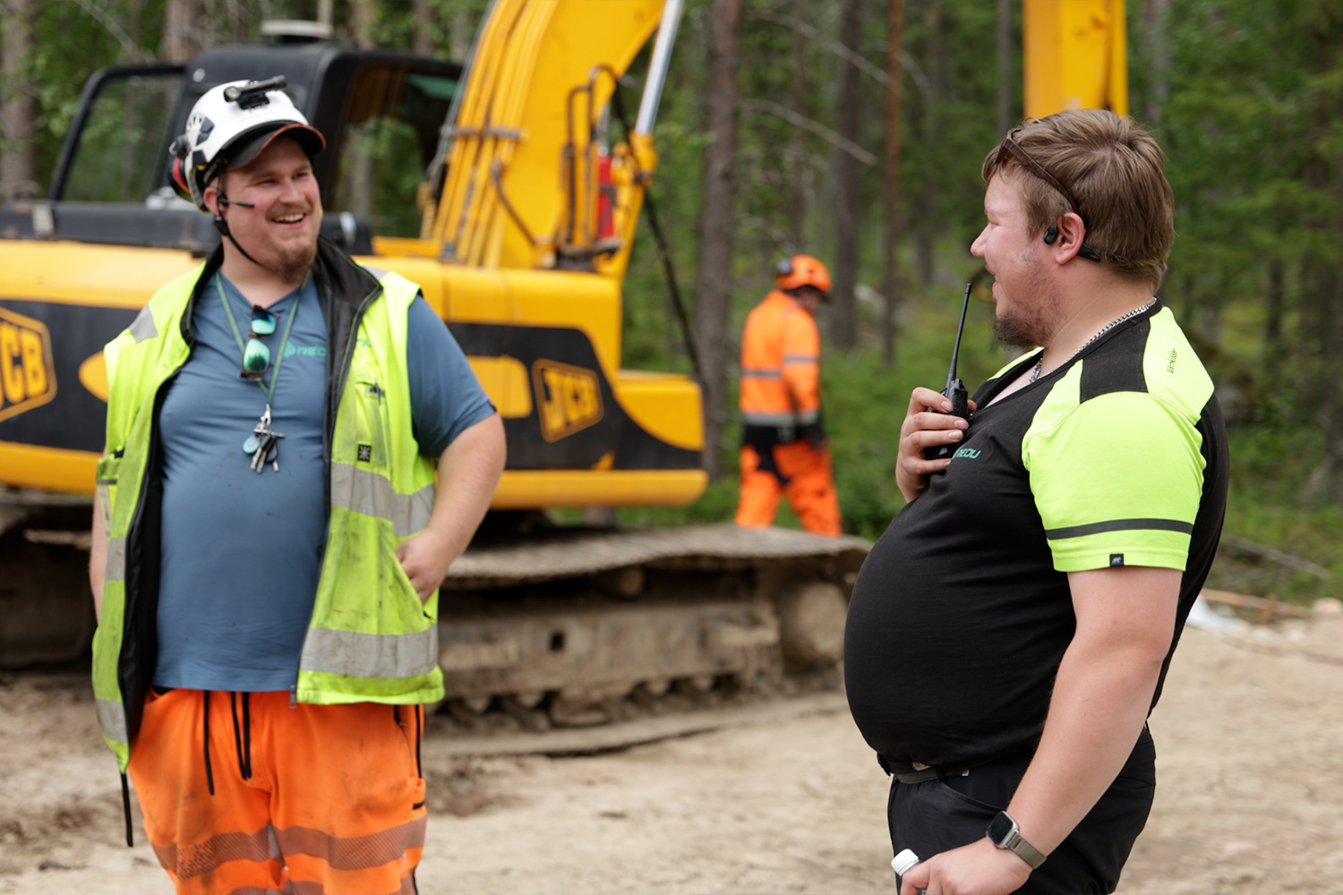 Kaksi maanrakennusalan opettajaa keskustelee työmaalla. Taustalla näkyy kaivinkone ja työmaa-alueella työskentelevä henkilö.