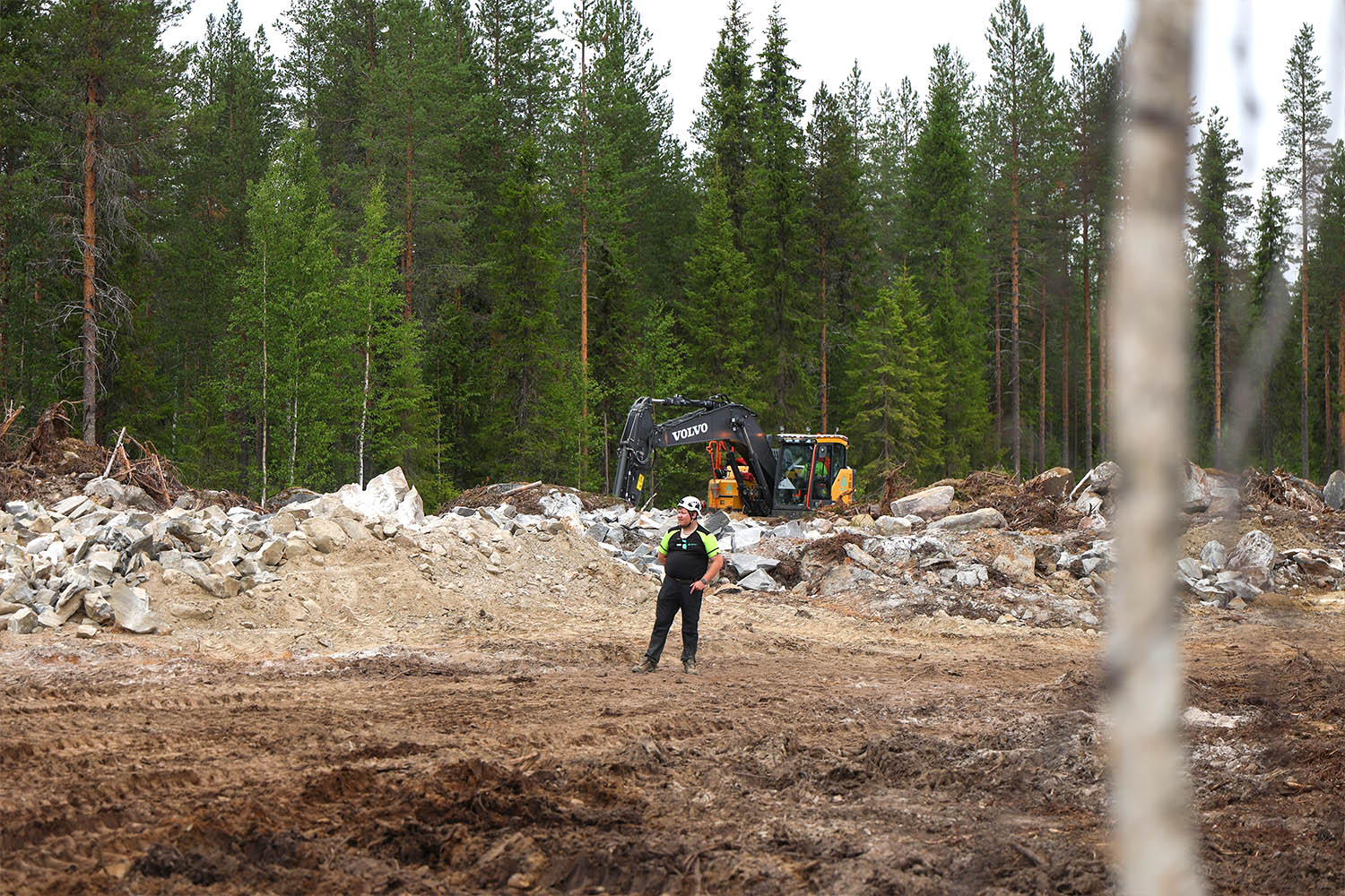Työmaa metsän reunassa, jossa henkilö seisoo avaralla maanrakennusalueella. Taustalla näkyy kaivinkoneita, louhetta ja metsämaisemaa.