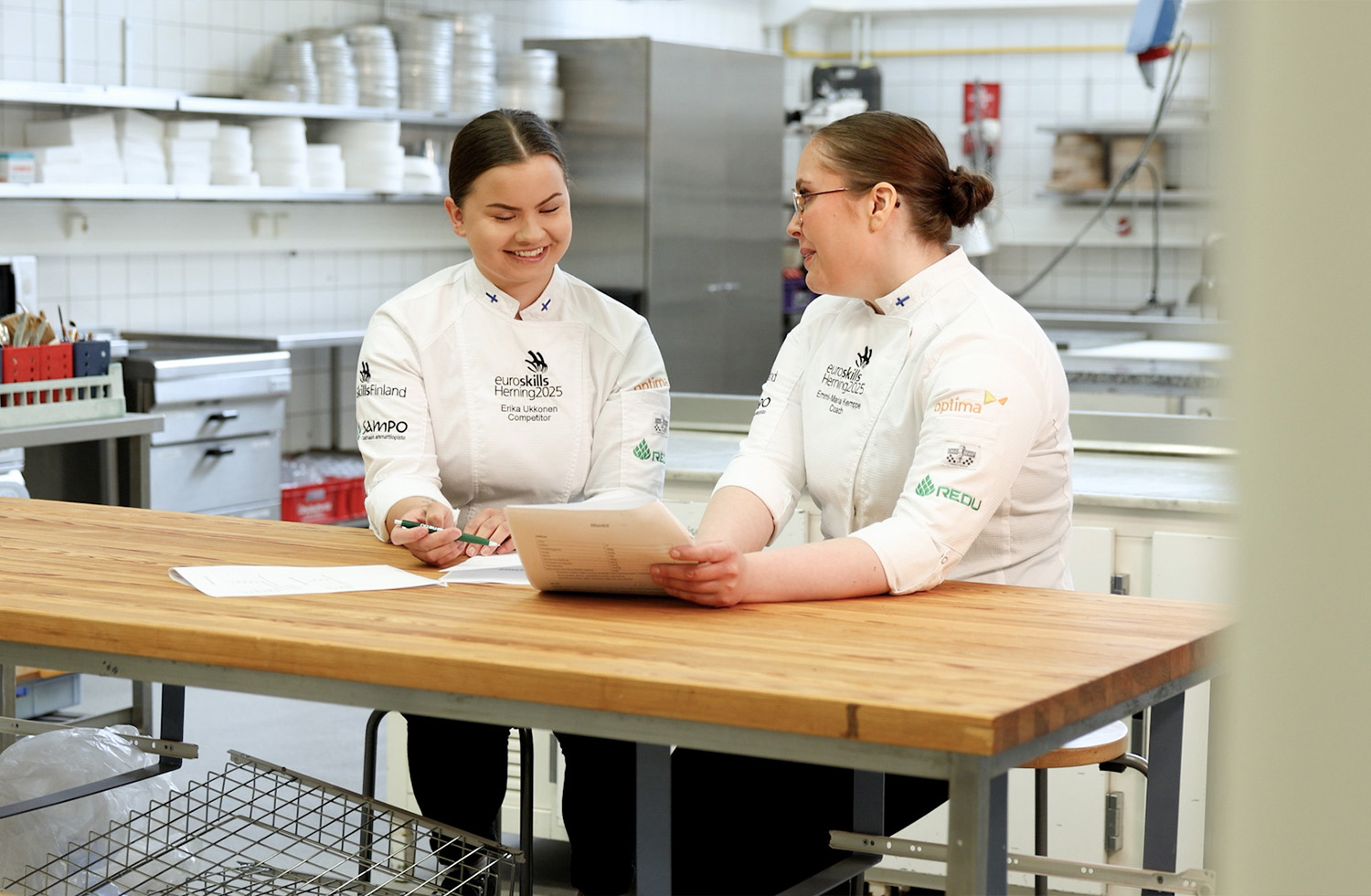 Emmi-Maria Kemppe ja Erika Ukkonen katsovat pöydän ääressä REDUn leipomolla papereita hymyillen.