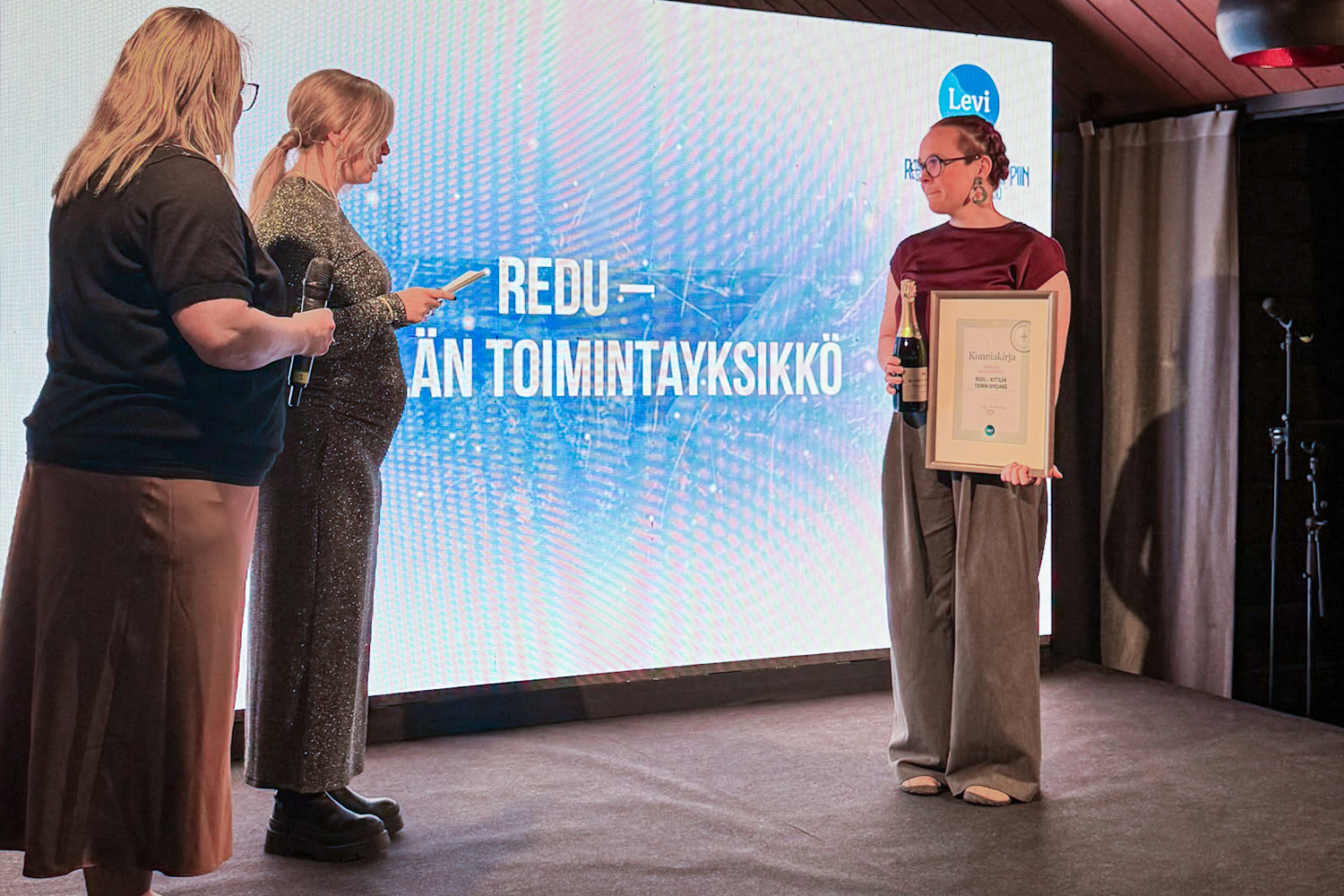 REDUn toimialapäällikkö Mirva Akkanen Levi Summitissa järjestetyssä Kipinäpäivien gaalassa vastaanottamassa Vuoden muu palveluyritys -tunnustuksen. Lavalla on kaksi tunnustuksen jakajaa lisäksi.