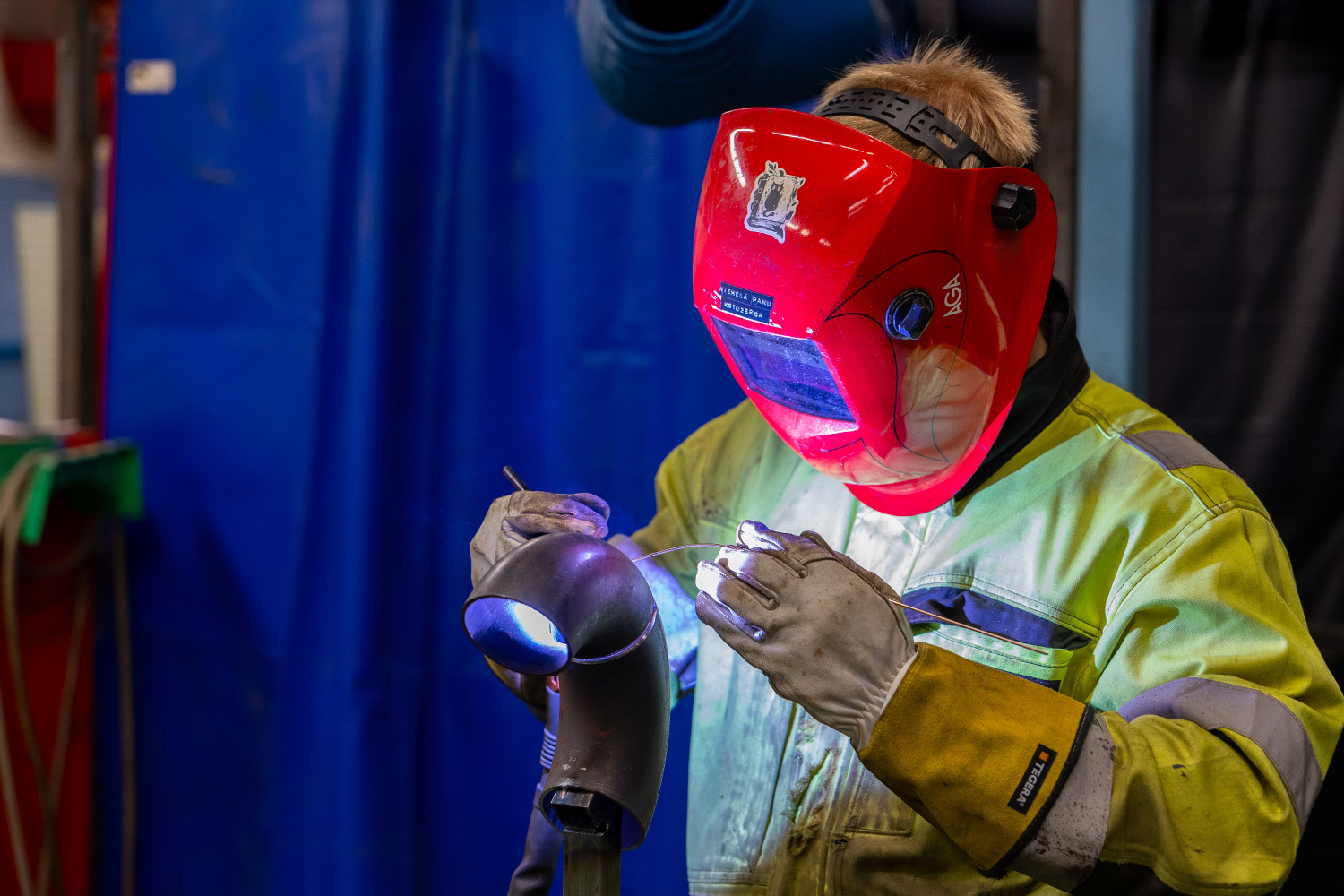 Henkilö punainen hitsausmaskin päässä hitsaamassa metallipalaa.