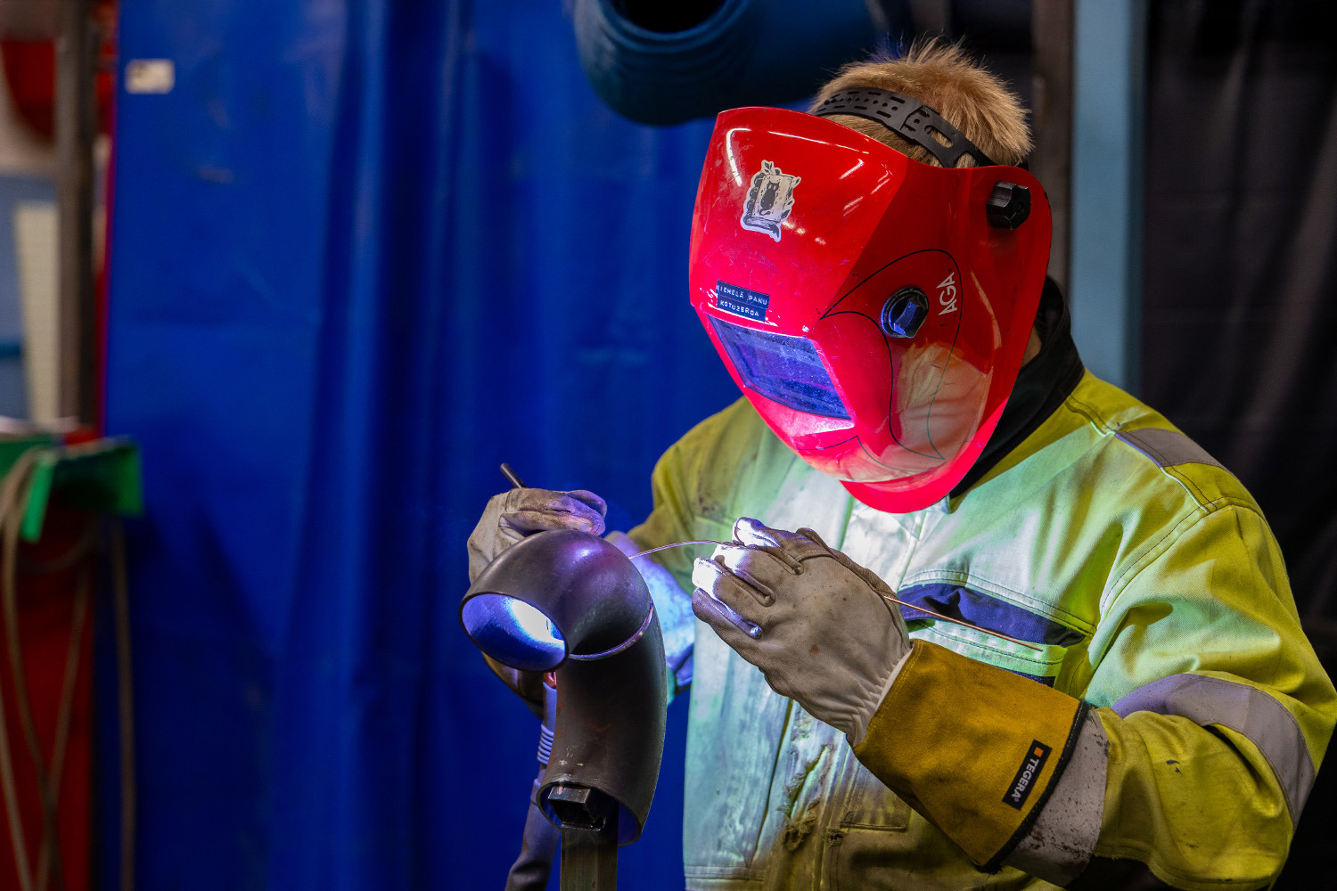 Henkilö punainen hitsausmaskin päässä hitsaamassa metallipalaa.